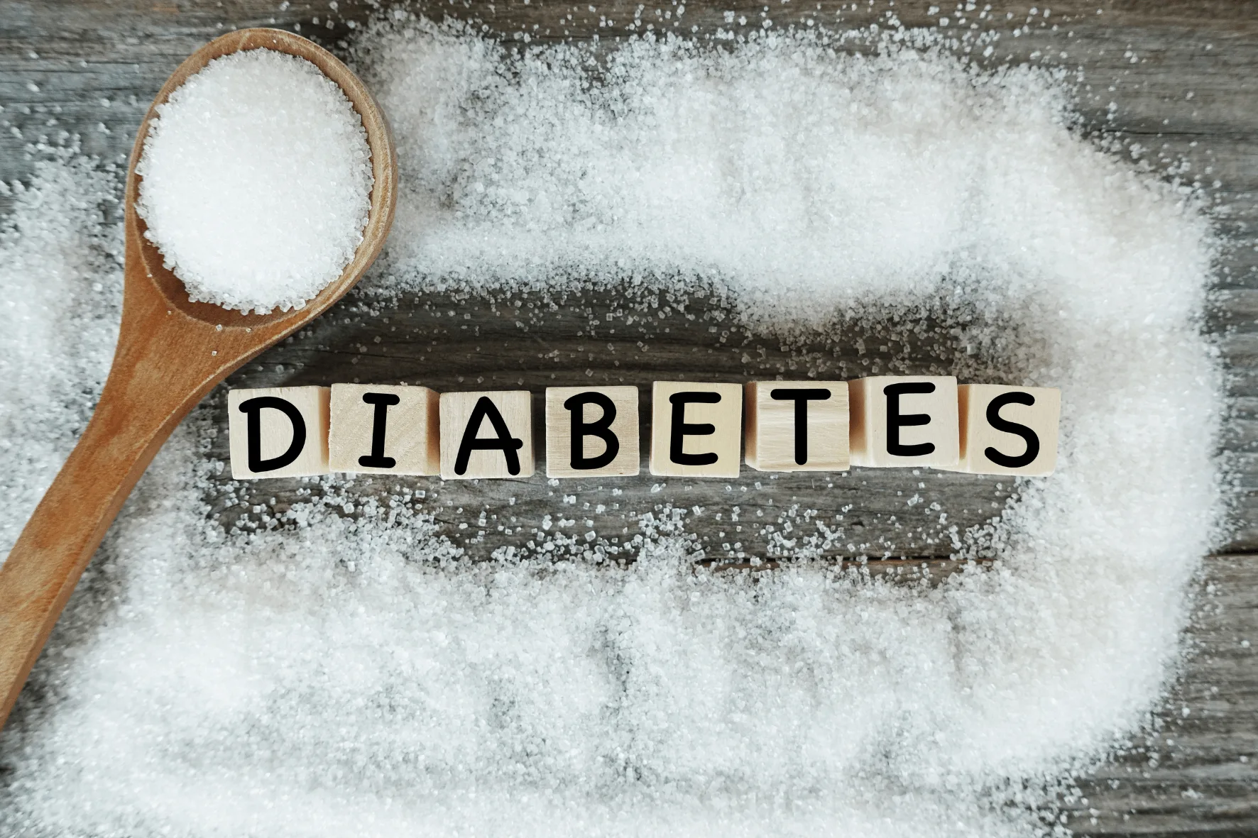 Blocos de madeira formando a palavra "diabetes" cercados de açúcar, com uma colher de açúcar ao lado